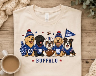 May include: A cream-colored t-shirt featuring a graphic of five dogs wearing blue and red sports jerseys. The dogs are depicted with football accessories including a foam finger, a helmet, a football, and a pennant flag that reads GO BILLS!
