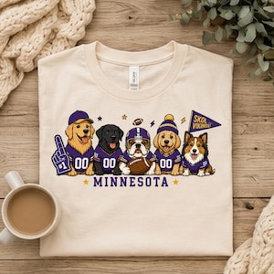 Peut inclure: Un t-shirt couleur crème avec un motif de cinq chiens portant des maillots de football violets et dorés. Les chiens sont représentés avec un doigt en mousse, un ballon de football et un fanion indiquant SKOL VIKINGS. Le texte MINNESOTA est imprimé en dessous.