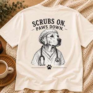 May include: A cream-colored short-sleeve t-shirt featuring a black ink-style illustration of a dog wearing a surgical cap and a stethoscope. The text "SCRUBS ON. PAWS DOWN." is printed above the dog, with a small paw print icon below the graphic.