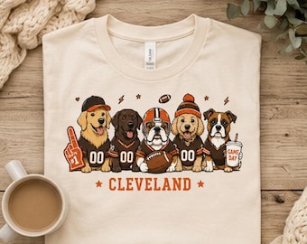 Puede incluir: Una camiseta de color crema con un gráfico de cinco perros con camisetas de fútbol americano marrones y naranjas. Los perros aparecen con un dedo de espuma, un balón y un vaso con la etiqueta GAME DAY. El texto CLEVELAND aparece debajo.