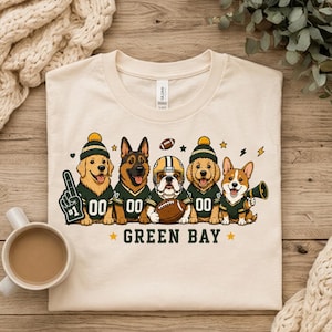 May include: A cream-coloured t-shirt featuring a graphic of five dogs wearing green and gold football jerseys and hats. The dogs are positioned above the text GREEN BAY. One dog holds a number one foam finger, and another holds a football.