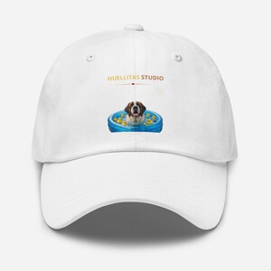 Puede incluir: Una gorra de béisbol blanca con un estampado gráfico de un perro marrón y blanco sentado en una piscina inflable azul llena de patitos de goma amarillos. Sobre el perro, el texto HUELLITAS STUDIO está impreso en naranja con un corazón rojo.