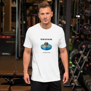 Puede incluir: Una camiseta blanca de manga corta con un gráfico de un perro en una pequeña piscina inflable azul llena de patitos de goma amarillos. El texto sobre el perro dice "TOO BIG FOR THIS POOL" y el texto debajo dice "too cute to care".