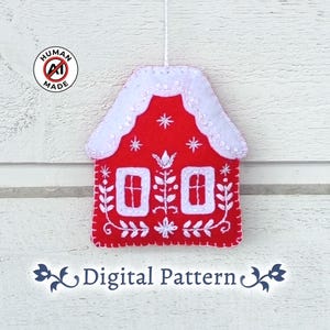 May include: A red felt house ornament featuring white embroidered floral details and two windows. The roof is covered in white felt with white stitching. A white string is attached to the top for hanging. The text "HUMAN MADE" and "Digital Pattern" appear.