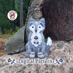 May include: A felt fox ornament in shades of grey, white, and navy blue, featuring intricate embroidery of a star on the forehead and floral motifs on the chest. The fox has a white tail tip and decorative stitching on its legs. Text reads Digital Pattern.