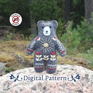 May include: A dark gray felt bear ornament featuring white, yellow, and red embroidered geometric patterns, a sun motif, and floral designs on the chest. The bear has a white face with black eyes and nose. The text Digital Pattern is visible.
