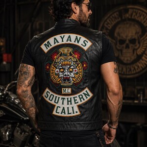 May include: A black leather vest featuring a large embroidered patch on the back. The patch displays a central Mayan-style mask graphic with the text MAYANS at the top and SOUTHERN CALI. M.C. at the bottom in green and white lettering.