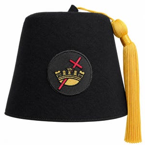 May include: A black felt fez hat featuring a circular embroidered patch on the front. The patch displays a gold crown positioned over a red cross. A long, bright yellow tassel is attached to the top center of the hat and hangs down the side.