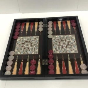 May include: A rectangular backgammon board featuring a dark frame and a central geometric inlay pattern. The board includes alternating red and light wood triangular points, with maroon and off-white circular game pieces arranged on the surface.