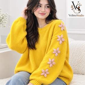 Könnte beinhalten: Ein leuchtend gelber, langärmeliger Strickpullover mit Rundhalsausschnitt und gerippten Bündchen. Der linke Ärmel ist mit fünf hellrosa, fünfblättrigen Blumenapplikationen in einer vertikalen Reihe verziert. Das VelinoStudio-Logo ist sichtbar.