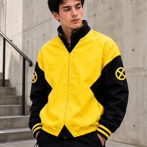 Young man standing by concrete stairs wearing yellow and black zip-up bomber jacket with X logo sleeve patch