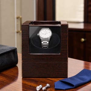 Könnte beinhalten: Ein dunkelbrauner Uhrenbeweger aus Holz mit einer klaren Glasfront, in dem eine silberfarbene Analoguhr mit weißem Zifferblatt und Metallgliederarmband auf einem schwarzen Samtkissen im quadratischen Sichtfenster präsentiert wird.