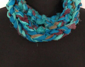 May include: A teal, turquoise, brown, and purple crocheted fabric necklace. The necklace is made of braided fabric strips and is thick and chunky.