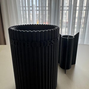 Puede incluir: Un recipiente cilíndrico negro con una textura exterior acanalada y el texto en relieve ART THO DESIGN en el lateral, acompañado de un inserto cilíndrico negro más pequeño con cuatro aletas perpendiculares en su cuerpo central.