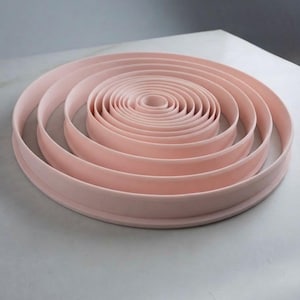 Puede incluir: Un conjunto de anillos circulares anidados en un suave color rosa pastel. Los anillos están dispuestos de forma concéntrica, con el anillo más pequeño en el centro y el más grande en el exterior, creando un patrón circular con bordes lisos.