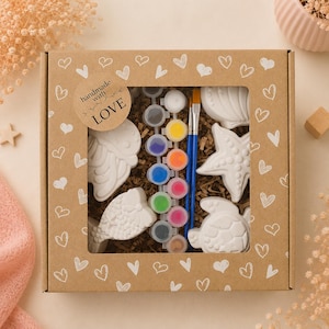 Könnte beinhalten: Ein quadratischer Bastelset-Karton mit Sichtfenster, verziert mit weißen Herzen und einem Aufkleber mit der Aufschrift "handmade with LOVE". Enthalten sind weiße Gipsfiguren mit Meeresmotiven, zehn kleine Farbtöpfe und zwei blaue Pinsel.