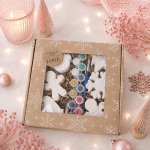 Può includere: Una scatola quadrata in cartone per kit creativi con motivo a fiocchi di neve e finestra trasparente. All'interno si trovano figure in gesso bianco, tra cui omini di pan di zenzero e fiocchi di neve, vasetti di vernice e un pennello.