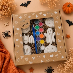 Può includere: Un kit creativo in una scatola di cartone marrone con finestra, contenente figure di Halloween in gesso bianco, tra cui un fantasma, zucche e un pipistrello. Il kit include due pennelli blu e una striscia di otto piccoli vasetti di vernice. La scatola presenta motivi a cuore bianchi e un adesivo con la scritta handmade with LOVE.