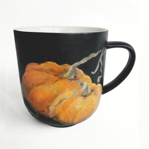Peut inclure: Une tasse en céramique noir mat ornée d'une citrouille orange peinte à la main avec des coups de pinceau visibles et une tige claire. L'intérieur de la tasse est blanc, tandis que l'extérieur est noir uni, créant un contraste avec la citrouille.