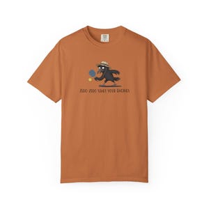 May include: A burnt orange short-sleeve t-shirt featuring a graphic of a black crow wearing a tan hat and holding a blue pickleball paddle. A yellow pickleball is nearby. The text below reads: ZERO-ZERO START YOUR ENGINES in black lettering.