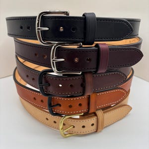 May include: A stack of five leather belts in varying shades of black, dark burgundy, dark brown, medium tan, and natural beige. Each belt features a single prong buckle, a matching leather keeper, and a series of punched holes for adjustment.
