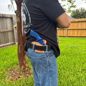 May include: A tan leather holster secured to a black belt, holding a bright blue training revolver. The holster features a visible logo stamp and is worn with blue denim jeans and a black t-shirt with white text on the back.