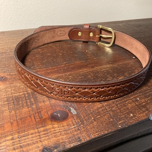 May include: A brown leather belt featuring a repeating diamond-shaped embossed pattern along the strap. The belt includes a brass-toned metal buckle and two brass rivets securing the leather loop. The letters SR are embossed on the leather surface.