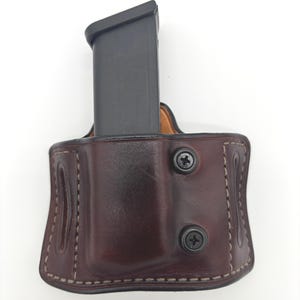 May include: A dark brown leather magazine pouch featuring visible stitching along the edges and two black circular screws on the front. A black rectangular firearm magazine is inserted into the top of the pouch, which is shaped to hold the item securely.
