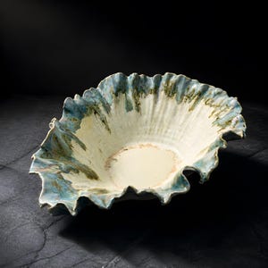May include: A ceramic bowl with a ruffled, shell-like edge featuring a cream-colored interior and a glaze that transitions into shades of blue and green along the textured, wavy rim, creating a distinct contrast against the dark background.