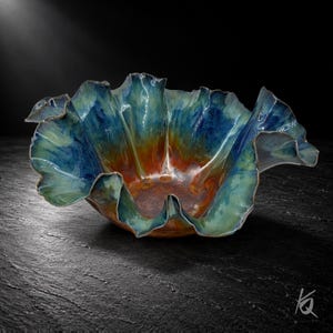 May include: A ceramic bowl with a ruffled, organic edge featuring a gradient glaze that transitions from a deep, rich copper-brown at the base to vibrant shades of teal, turquoise, and cobalt blue along the undulating, flared rim.