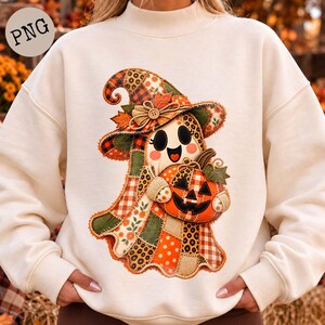 May include: A cream-coloured long-sleeve sweatshirt featuring a central graphic of a ghost wearing a patchwork witch hat and holding a jack-o'-lantern. The ghost design incorporates various patterns including plaid, leopard print, and floral motifs in orange, green, and brown tones.