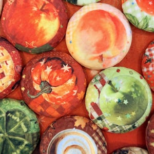 May include: A collection of circular fabric-covered buttons featuring various printed designs including a red apple, a peach, a pumpkin, a green apple, a kiwi slice, and a coffee latte swirl, all arranged closely together on an orange fabric surface.