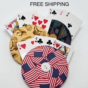 May include: Three circular fabric-covered buttons featuring distinct patterns: one with tan cowboy boots, one with a dark geometric print, and one with an American flag design. Each button is partially tucked behind standard playing cards.