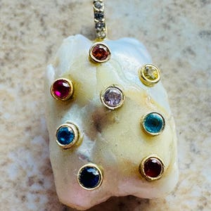 May include: A baroque pearl pendant featuring a gold bail set with small clear stones. The pearl surface is adorned with seven bezel-set gemstones in various colors, including red, yellow, light purple, blue, dark blue, and deep red hues.