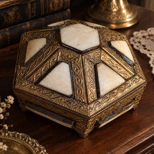 Pode incluir: Uma caixa hexagonal com uma tampa que apresenta uma incrustação central de pedra branca rodeada por seis painéis menores de pedra branca. A estrutura é feita de metal dourado texturizado com padrões intrincados ao longo das bordas.