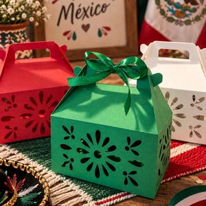Puede incluir: Una caja de regalo de papel verde con un patrón floral troquelado y un lazo de cinta de satén verde en la parte superior. La caja tiene un asa y forma parte de un conjunto que incluye una caja roja y una blanca, con diseños y cortes a juego.
