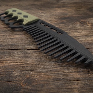 May include: A matte black hair comb designed with a tactical knife aesthetic, featuring a pointed tip, a series of rectangular cutouts along the spine, and a sage green handle grip accented with three small black diamond-shaped inlays.