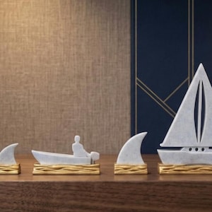 May include: A collection of six nautical-themed figurines crafted from light gray stone with gold-painted wave-textured bases. The set includes a canoe with a person, two shark fins, a motorboat with a person, and a sailboat with a triangular sail.