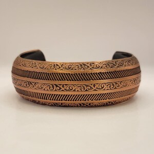 May include: A copper bracelet with an intricate engraved design featuring a repeating pattern of swirls and lines.