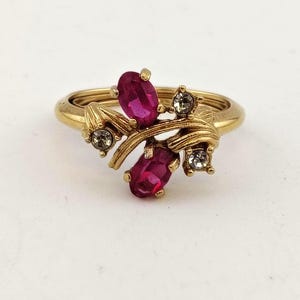 May include: A gold-toned ring featuring a floral design with two oval-cut, magenta-colored gemstones and four smaller, clear gemstones. The ring has a delicate, intertwined band and is set against a white background.