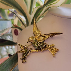 May include: Gold-tone brooch featuring two hummingbirds with green rhinestone accents. The larger hummingbird is perched on a branch with its wings spread out. The smaller hummingbird is perched on the branch below the larger hummingbird.