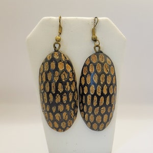 May include: A pair of oval-shaped earrings with a black and gold pattern. The earrings are made of wood and have a unique, textured surface.