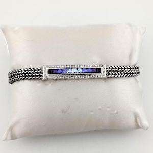 K&ouml;nnte beinhalten: Ein silbernes Armband mit einem rechteckigen Mittelst&uuml;ck mit einem Farbverlauf aus blauen Edelsteinen. Das Mittelst&uuml;ck ist mit kleinen, klaren Steinen eingefasst. Das Armband ist ein gewebtes Design in Schwarz und Silber.