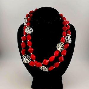 May include: A two-strand necklace featuring vibrant red, irregularly shaped beads. The necklace is accented with silver-colored, ornate circular and cylindrical beads. The necklace is displayed on a black mannequin bust against a white background.