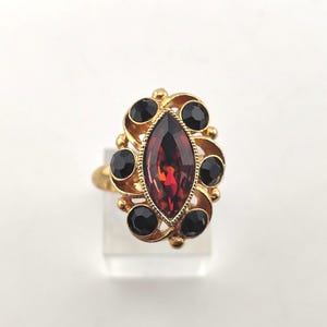 May include: A gold-toned ring featuring a central marquise-cut deep red gemstone surrounded by six smaller round black stones. The setting has a decorative, swirling metal frame that encircles the central stone and holds the black accents.