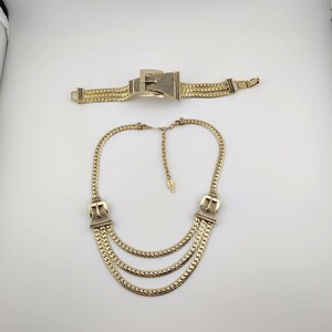 May include: A gold-coloured jewellery set comprising a necklace and bracelet. The necklace has three strands and buckle-style clasps. The bracelet has a similar design with a buckle clasp. Both pieces are made of textured metal.