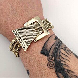 May include: A gold-coloured bracelet with a buckle design. The bracelet features a large, textured buckle with a rectangular frame and a pin closure. The band is made of multiple strands of gold-coloured metal, creating a bold, statement piece.