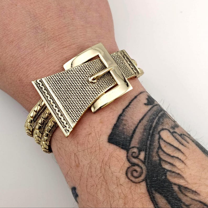 May include: A gold-coloured bracelet with a buckle design. The bracelet features a large, textured buckle with a rectangular frame and a pin closure. The band is made of multiple strands of gold-coloured metal, creating a bold, statement piece.