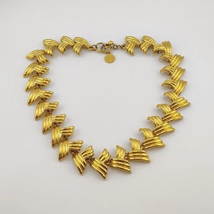 May include: A gold-toned necklace with a repeating geometric design. The necklace features a series of stylized, layered shapes connected by a delicate chain and clasp. The jewelry is displayed against a white background.