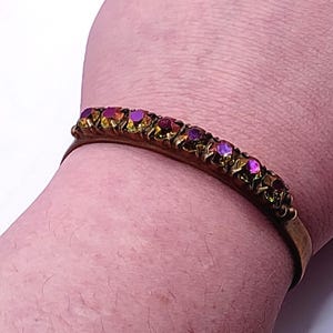 May include: A vintage gold-coloured metal bangle featuring a line of iridescent, multi-coloured gemstones. The gemstones display hues of purple, red, and gold. This classic bangle is an elegant piece of jewellery.